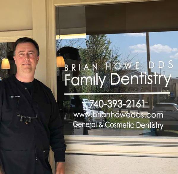 The image shows a man standing in front of a sign advertising Brian Howe DDS Family Dentistry with a phone number and website provided, indicating that he is likely an employee or owner of the dental practice.