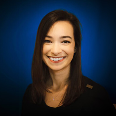 The image shows a woman smiling at the camera. She has short brown hair and is wearing a black top with a necklace. Behind her, there is a blue background with a gradient effect.