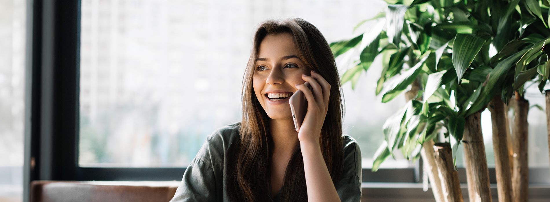 A young woman with long hair, wearing a green top, sitting at a table, smiling and talking on her cell phone while seated indoors with potted plants in the background.